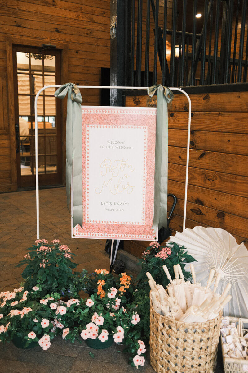 Wedding welcome sign with pink border stands indoors, surrounded by potted pink-and-white flowers and a basket of paper parasols. Perfect for a colorful summer wedding, the sign reads, “Welcome to our wedding, Peyton & Milo,” adding personal touches to your special day.