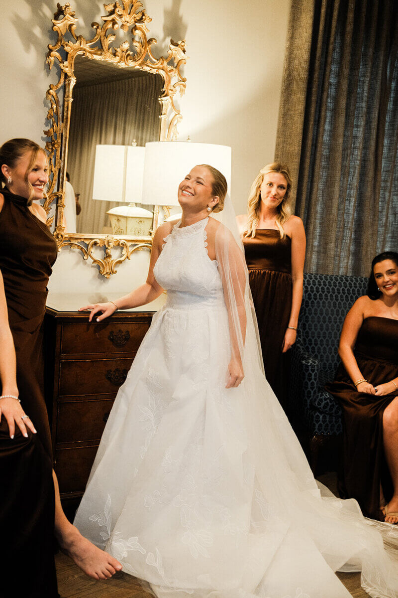 A bride in a white gown stands smiling in front of a mirror, surrounded by bridesmaids in brown dresses, adding personal touches that make this colorful summer wedding truly unforgettable.