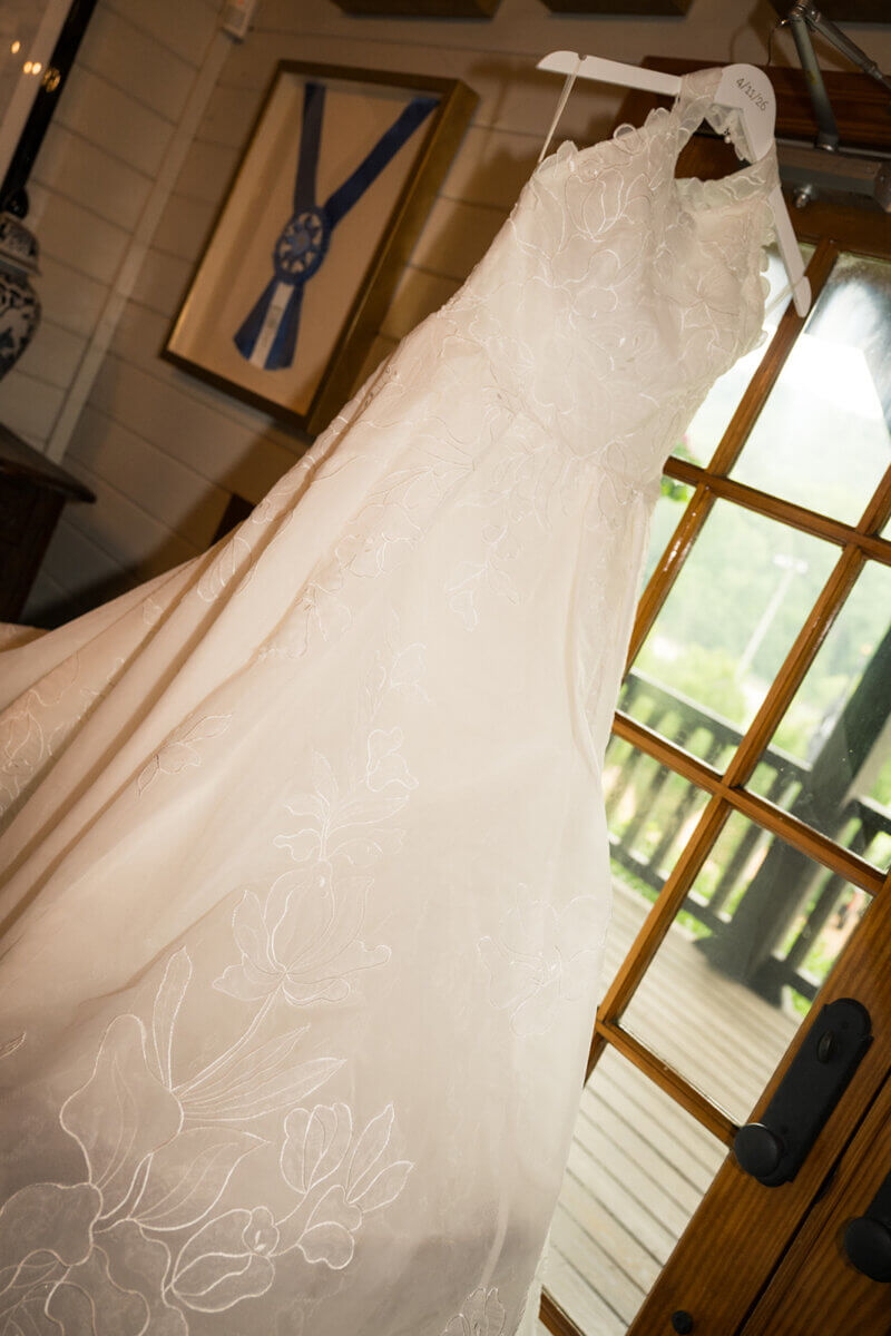 A white lace wedding dress hangs on a personalized hanger by a glass door, with natural light illuminating its floral details—a perfect reflection of the personal touches that make a summer wedding truly unforgettable.