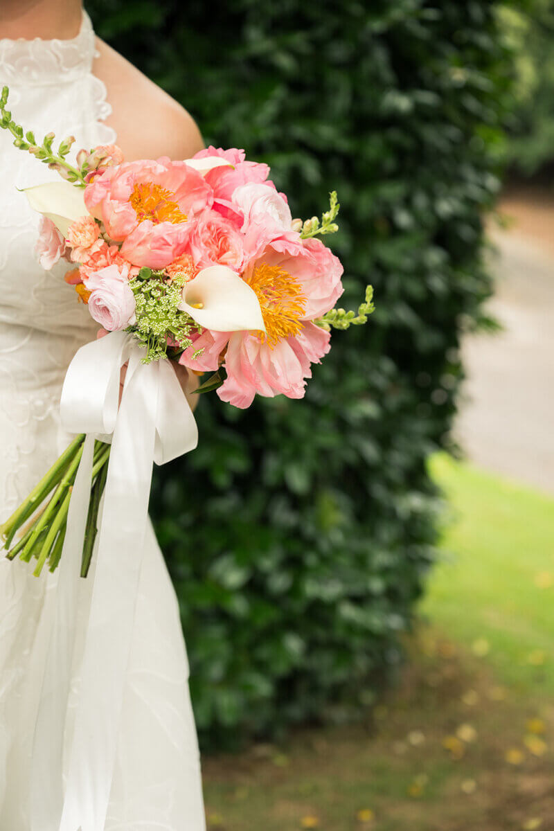 Person in a white dress holds a bouquet of pink peonies, white calla lilies, and greenery, tied with a white ribbon, standing outdoors near green foliage—a perfect scene for a colorful summer wedding with personal touches reflected in every detail.