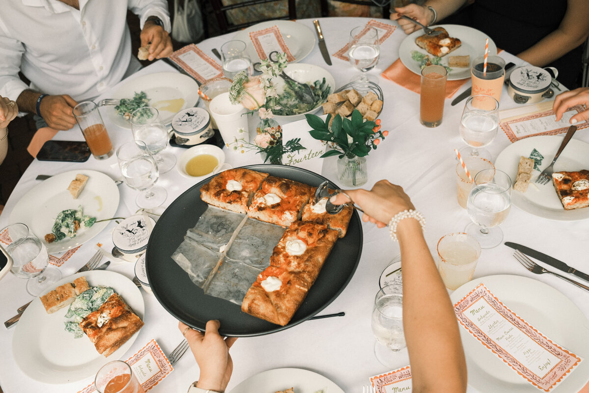 People seated around a table with various foods—including rectangular pizza slices being served, plates, drinks, menus, and a salad centerpiece—enjoy a meal enhanced by personal touches that make the gathering feel warm and inviting.