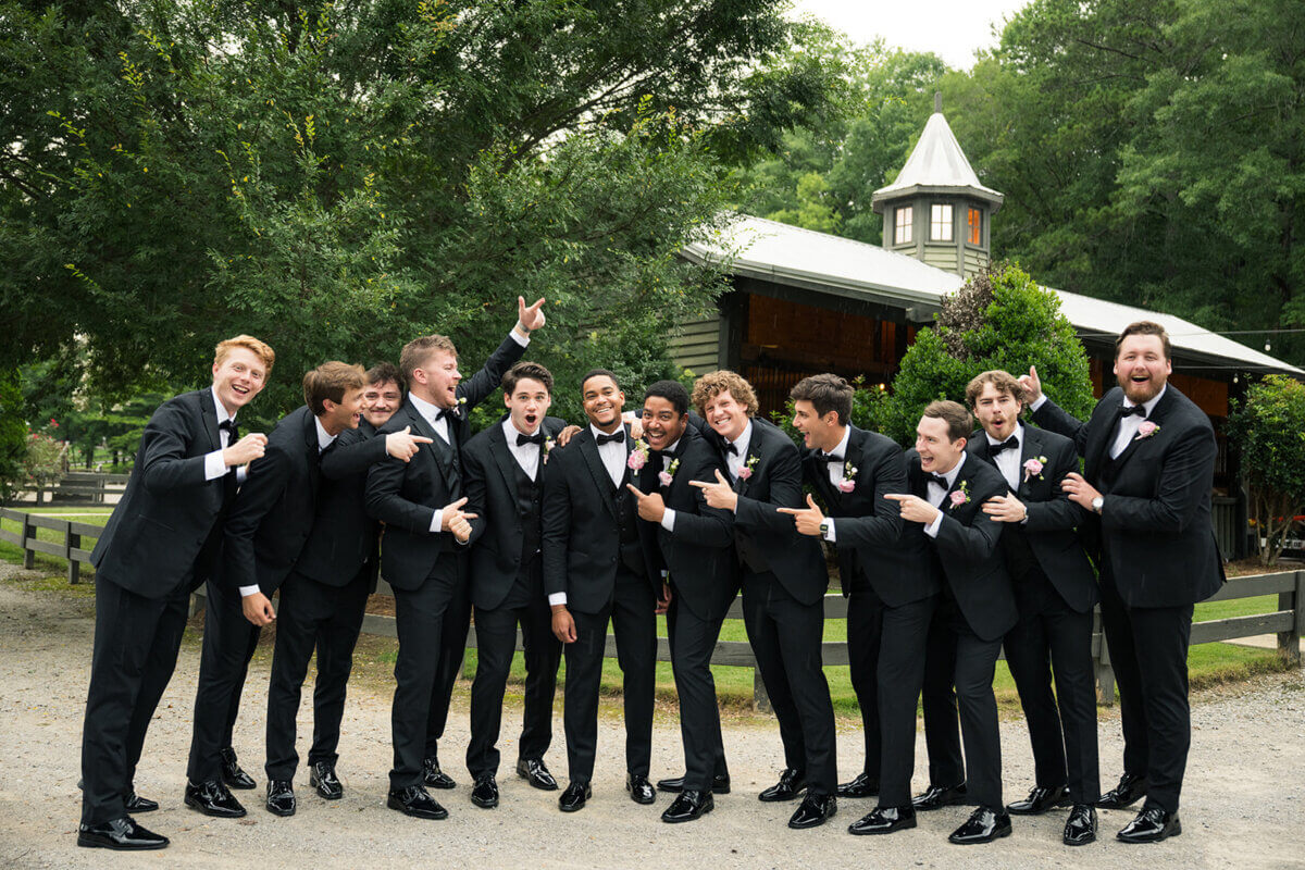 Twelve men in black suits and bow ties stand outdoors, smiling and pointing at the groom, adding personal touches to this colorful summer wedding with a barn and trees in the background.