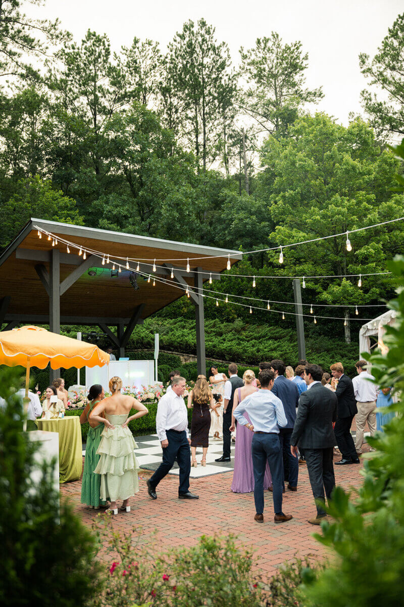 People in formal attire socialize outdoors under string lights near a pavilion surrounded by trees and greenery, creating an inviting atmosphere for a colorful summer wedding filled with personal touches.