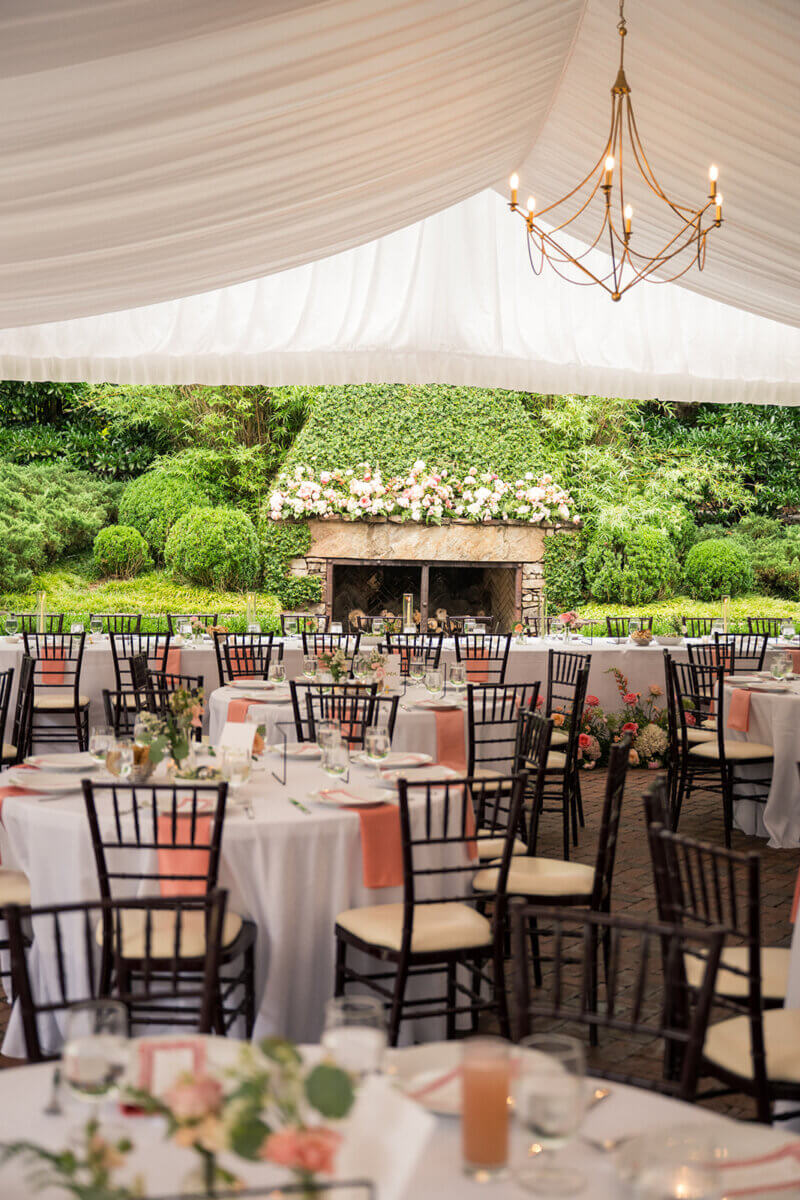 Outdoor wedding reception setup with round tables covered in white linens, dark chairs, peach accents, and a floral-decorated fireplace under a white draped tent with a chandelier—perfect for a Colorful Summer Wedding filled with personal touches.