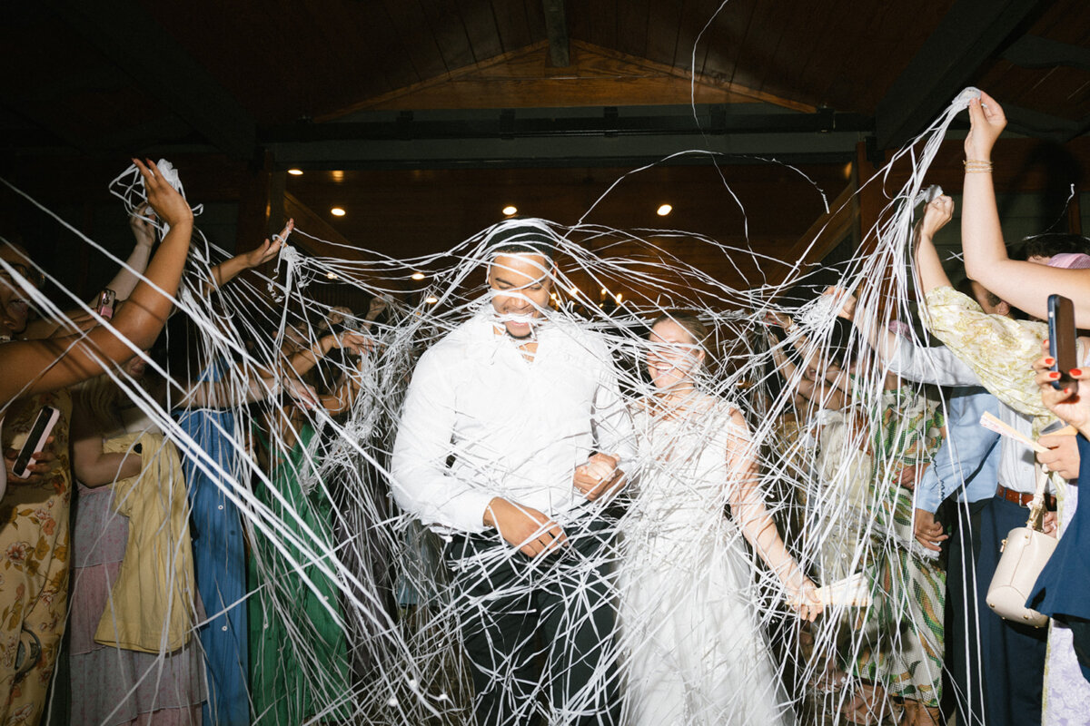 A couple walks through a crowd as guests spray them with white string; everyone appears to be celebrating indoors under warm lighting, with personal touches making the joyful moment truly memorable.