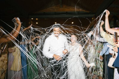 A couple walks through a crowd as guests spray them with white string; everyone appears to be celebrating indoors under warm lighting, with personal touches making the joyful moment truly memorable.