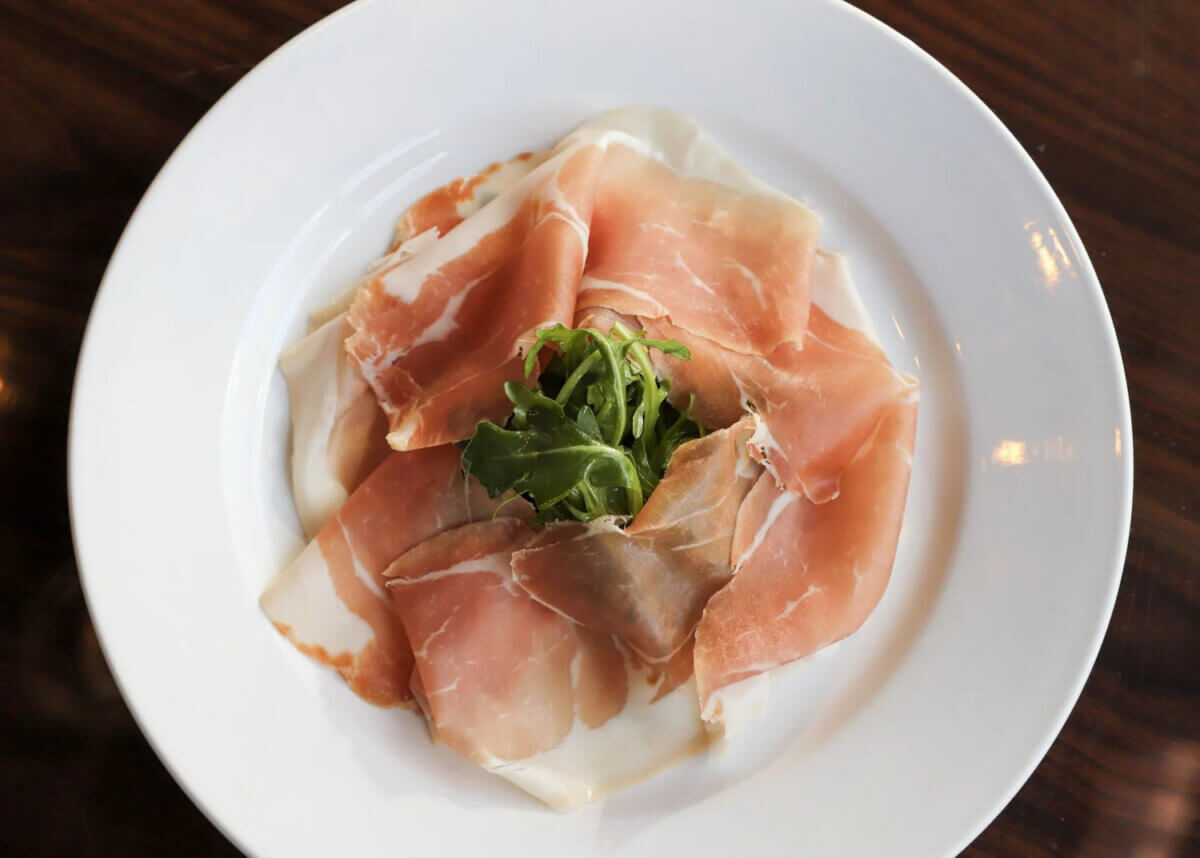 Thin slices of cured meat arranged on a white plate with a small pile of arugula in the center, set on a dark wooden table—this dish brings the flavors of international cuisine to Birmingham, letting you eat your way around the world without leaving the city.