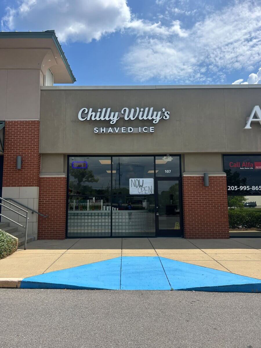 Front view of Chilly Willy’s Shaved Ice store—one of the refreshing new restaurants in Birmingham dining—features a handwritten “Now Open” sign in the window, a blue curb ramp in the foreground, and a partly cloudy sky above.