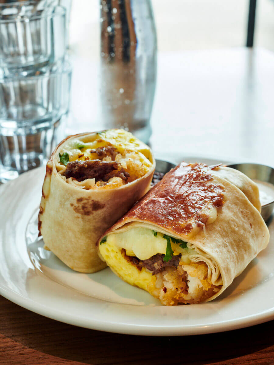 A breakfast burrito cut in half on a white plate, showing eggs, sausage, cheese, and vegetables inside—a delicious example of the creative morning fare served at new restaurants in Birmingham. A metal cup and stacked glasses are in the background.