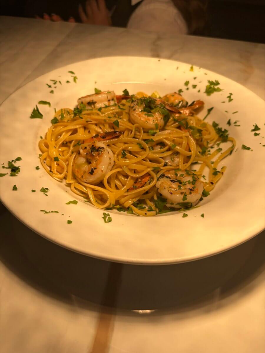 A white bowl of linguine pasta with shrimp, garnished with chopped parsley, sits on a marble surface under warm lighting—making it a tempting choice among the standout dishes served at new restaurants in Birmingham.