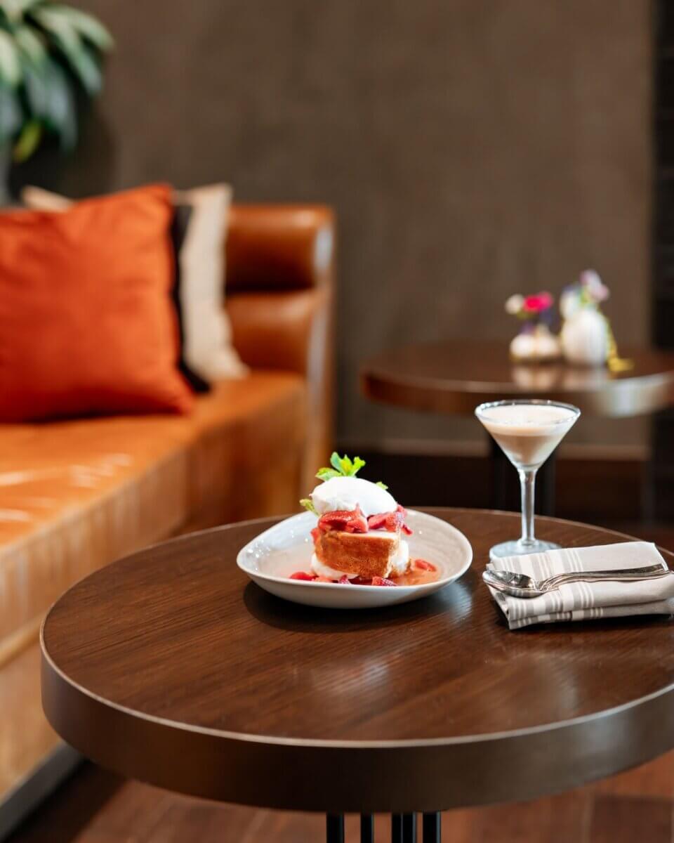 A dessert with whipped cream and sauce sits on a plate next to a cocktail on a round wooden table, with napkins and a fork in a modern lounge setting—an inviting scene that captures the elevated ambiance of Birmingham dining.