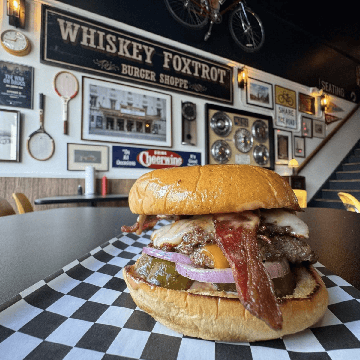 A cheeseburger with bacon, onions, pickles, and sauce sits on a checkered paper in a diner decorated with vintage signs and memorabilia—one of the must-try places to eat among Birmingham restaurants.