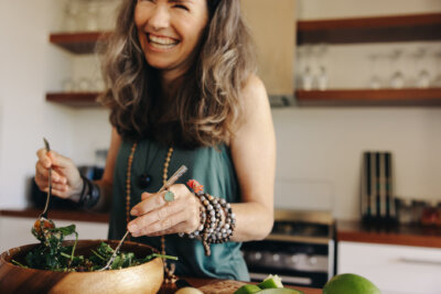 Woman in sleeveless top smiles while mixing a salad in a wooden bowl in a modern kitchen, embracing the principles of the longevity diet and showing how eating for longevity can add years to life.
