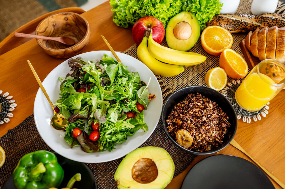 A table with a green salad, granola, avocado, bananas, apples, orange slices, lettuce, green bell pepper, bread, and a glass of orange juice—perfect for those interested in eating for longevity and choosing foods that may add years to your life.