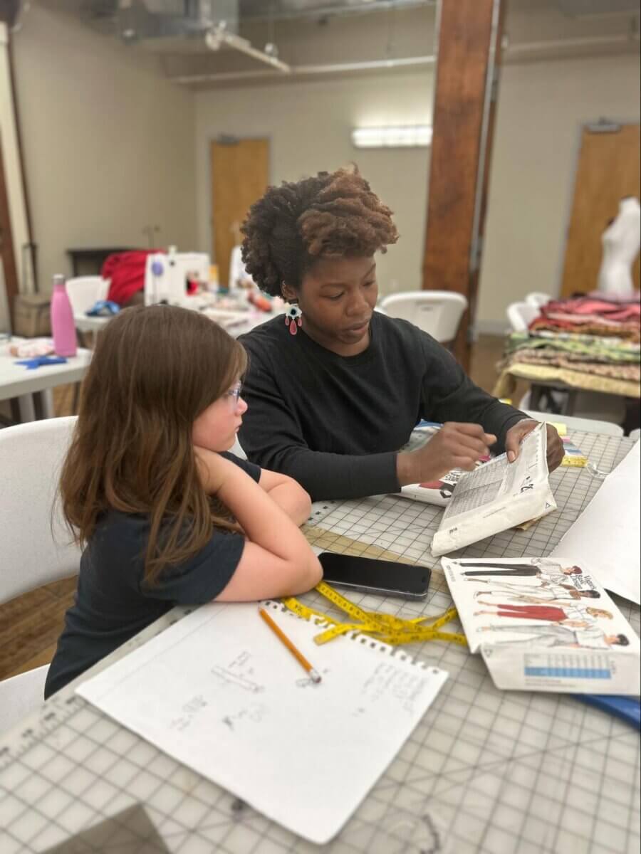 She Brings Bold Fashion, Cultural Heritage & Creativity to Memphis - 6 An adult and a child sit at a table with fabric and sewing materials, looking at a sewing pattern booklet and notes.
