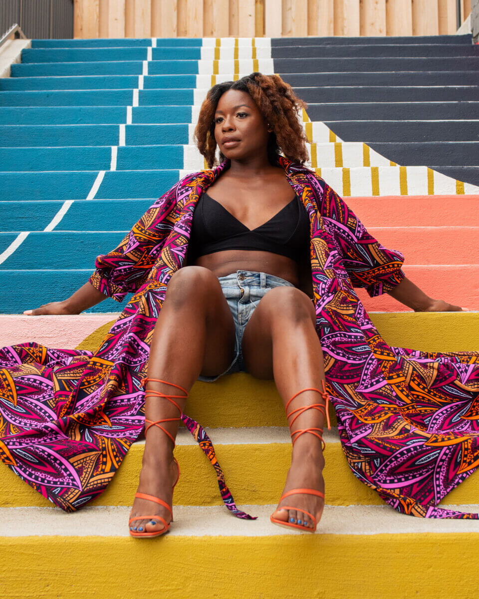 She Brings Bold Fashion, Cultural Heritage & Creativity to Memphis - 4 Woman in a colorful robe, black top, and denim shorts sits on multicolored striped stairs, looking to the side with legs extended.