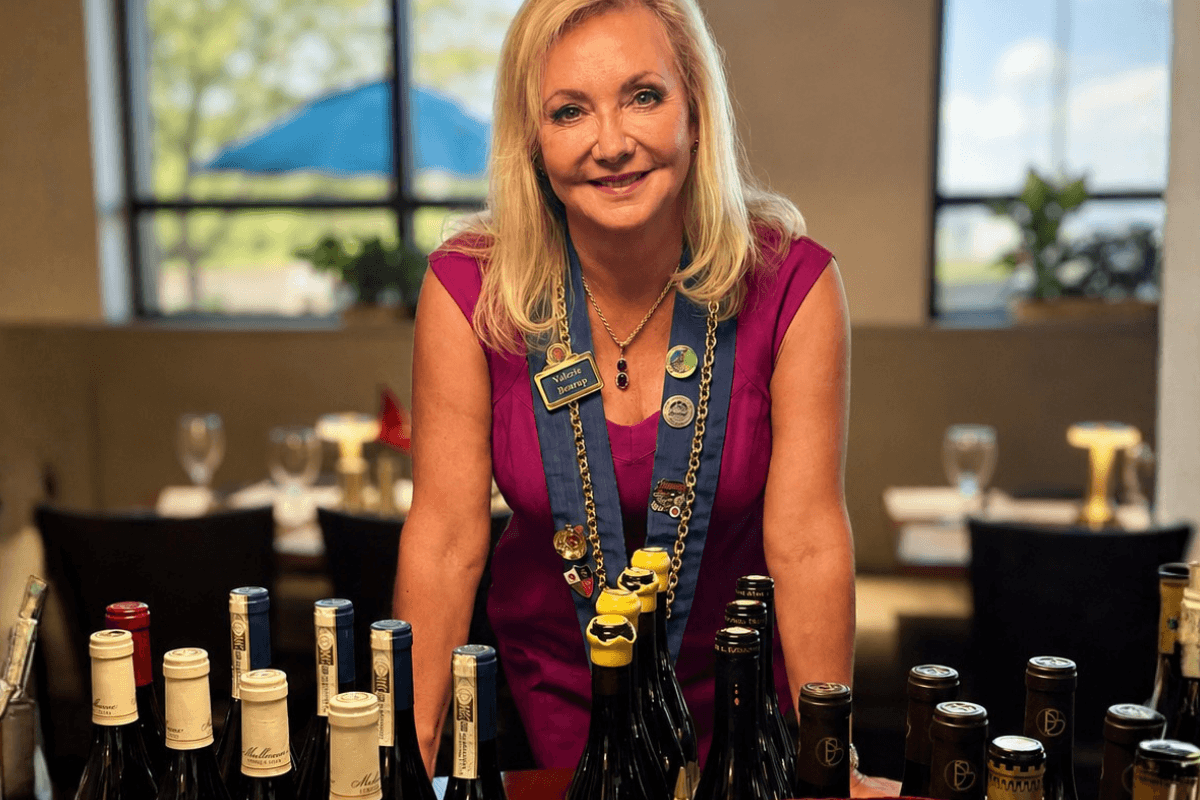 A woman in a purple dress stands behind a table with assorted wine bottles in a well-lit dining room.