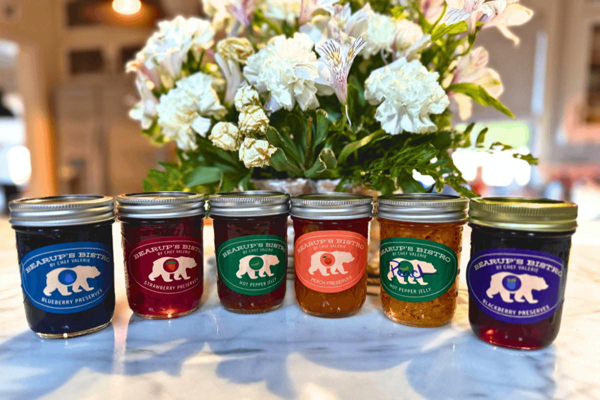 Six jars of Bearup’s Bistro preserves and jellies are lined up on a counter, with assorted flavors and colored labels, in front of a bouquet of white flowers.