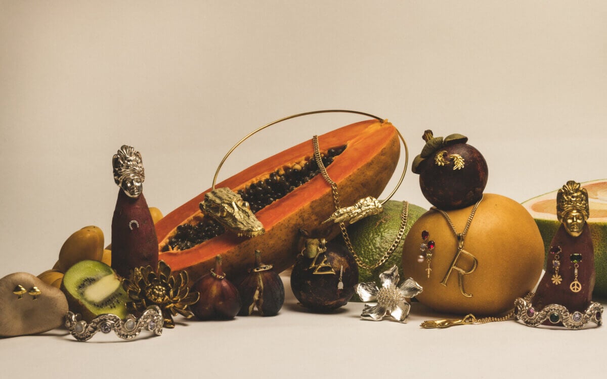 Gators, Magnolias, and Unicorn Horn Shells: Meet the New Orleans Designer Turning Southern Nature Into Jewelry - 3 Assorted gold and silver jewelry displayed on and around sliced papaya, mango, kiwi, and other fruits against a plain background.
