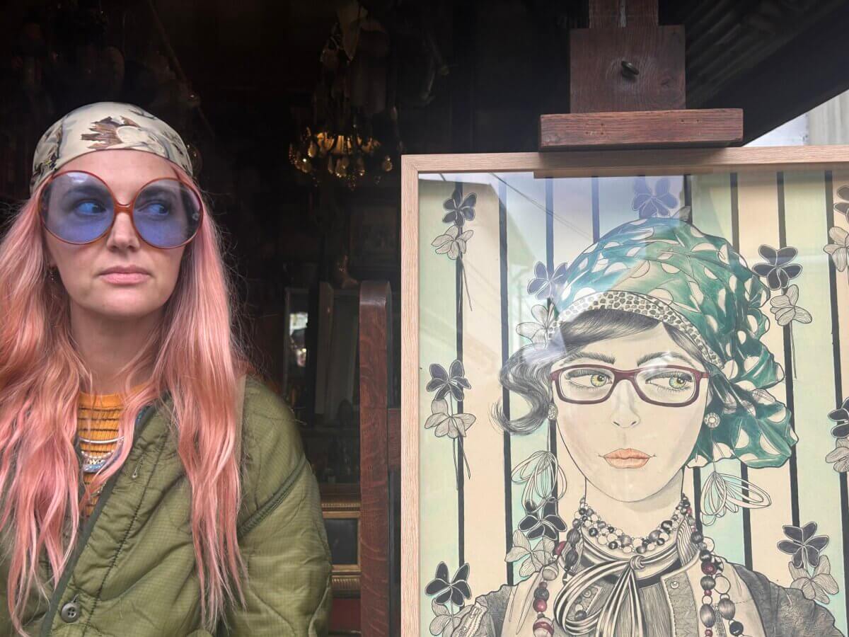 Gators, Magnolias, and Unicorn Horn Shells: Meet the New Orleans Designer Turning Southern Nature Into Jewelry - 2 A woman with pink hair, blue sunglasses, and a headscarf stands next to a framed illustration of a similarly styled woman.