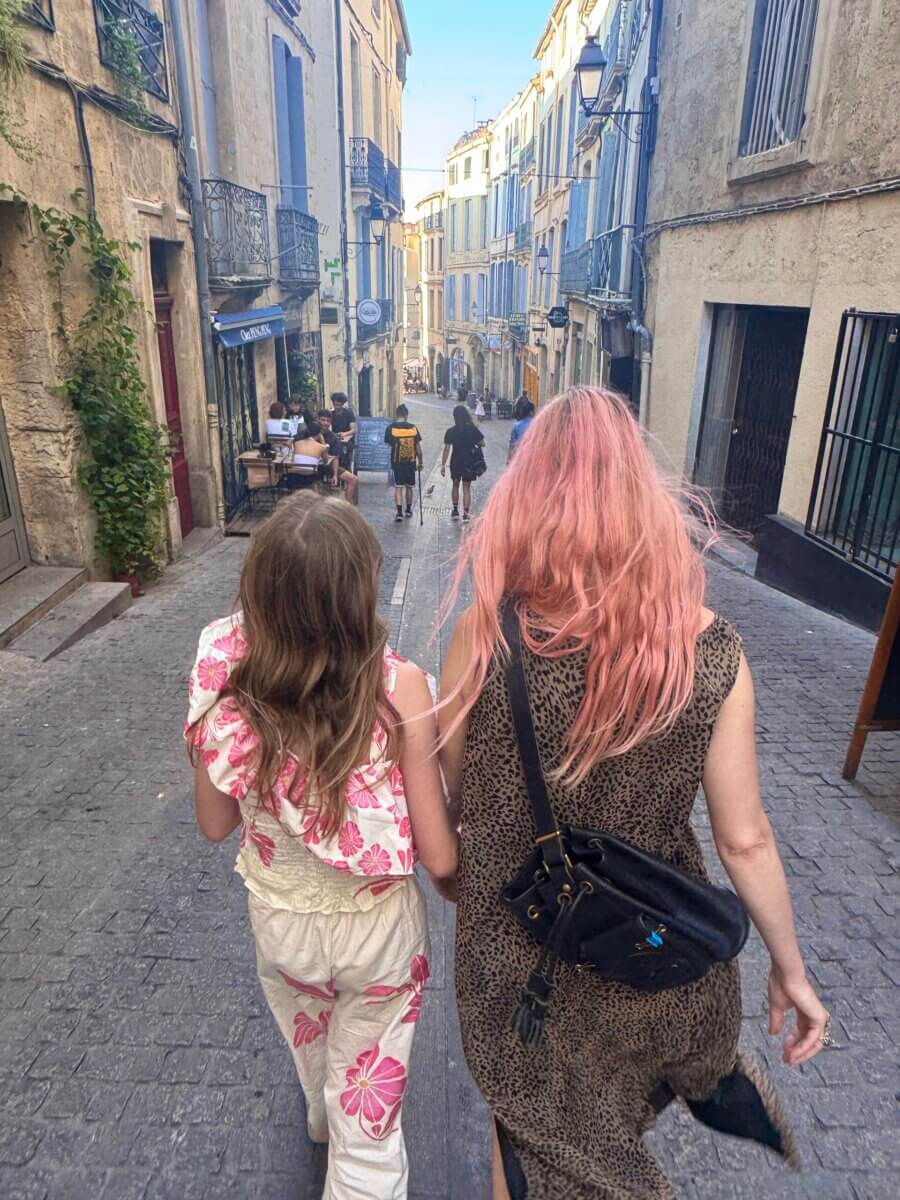 Gators, Magnolias, and Unicorn Horn Shells: Meet the New Orleans Designer Turning Southern Nature Into Jewelry - 6 Two people with long hair, one with pink hair, walk down a narrow, cobblestone street lined with old buildings. Other people are seen further ahead on the street.