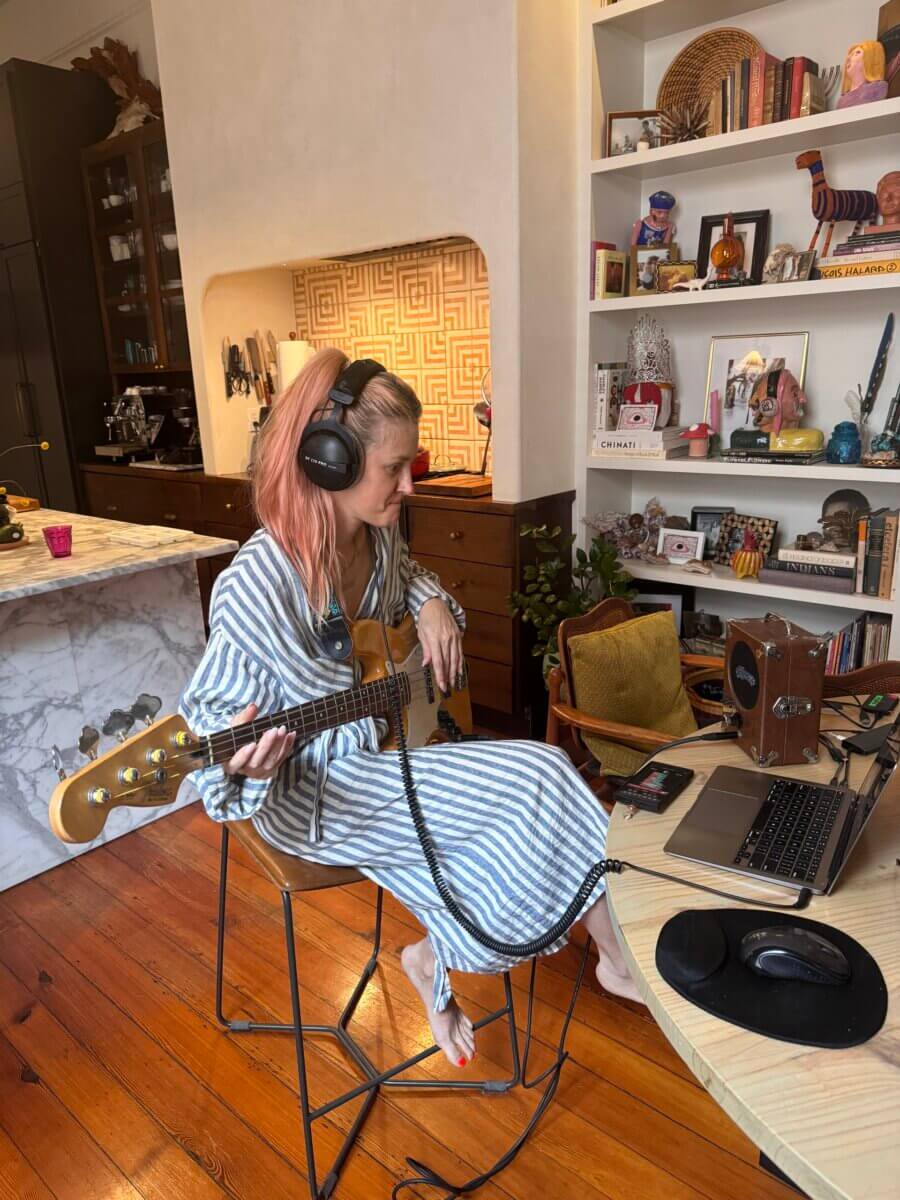 Gators, Magnolias, and Unicorn Horn Shells: Meet the New Orleans Designer Turning Southern Nature Into Jewelry - 5 A woman with headphones and a striped dress plays an electric bass guitar while sitting in front of a laptop and speakers in a cozy, book-filled home workspace.
