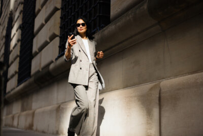 A person in a light gray suit and sunglasses walks outside next to a stone building on a sunny day, channeling Fall Fashion 2026 with a smartphone in hand.