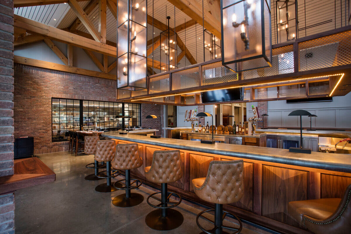 Lake Oconee: Your Guide to a Luxe Georgia Getaway - 3 Modern bar interior with leather barstools, wooden countertops, exposed beams, brick walls, pendant lighting, and a glass window showing a dining area in the background—an inviting spot perfect for your Lake Oconee weekend or as a stylish retreat during your next visit to Greensboro GA.