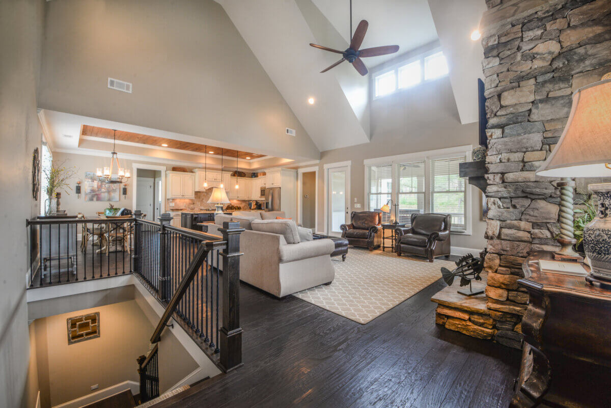 Lake Oconee: Your Guide to a Luxe Georgia Getaway - 2 Open-concept living area with vaulted ceiling, stone fireplace, kitchen, dining table, large windows, and staircase leading downstairs. Neutral tones and modern furnishings throughout create a welcoming retreat in Greensboro GA—perfect for relaxing after a day spent exploring Lake Oconee.
