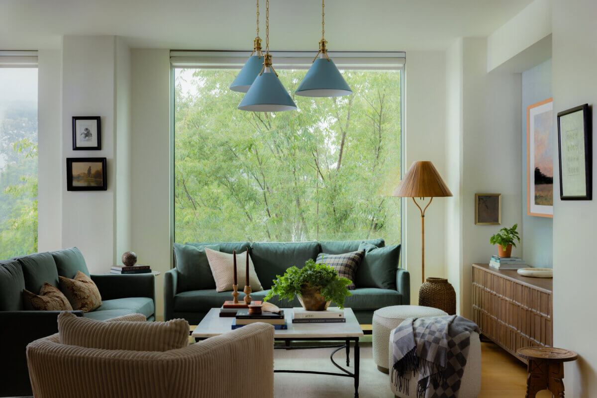 Whimsy Meets Cozy in This Dreamy Carolina Blue Home - 2 Modern living room with large windows, green sofas, a wooden coffee table, indoor plants, and blue pendant lights. Trees are visible outside through the windows.