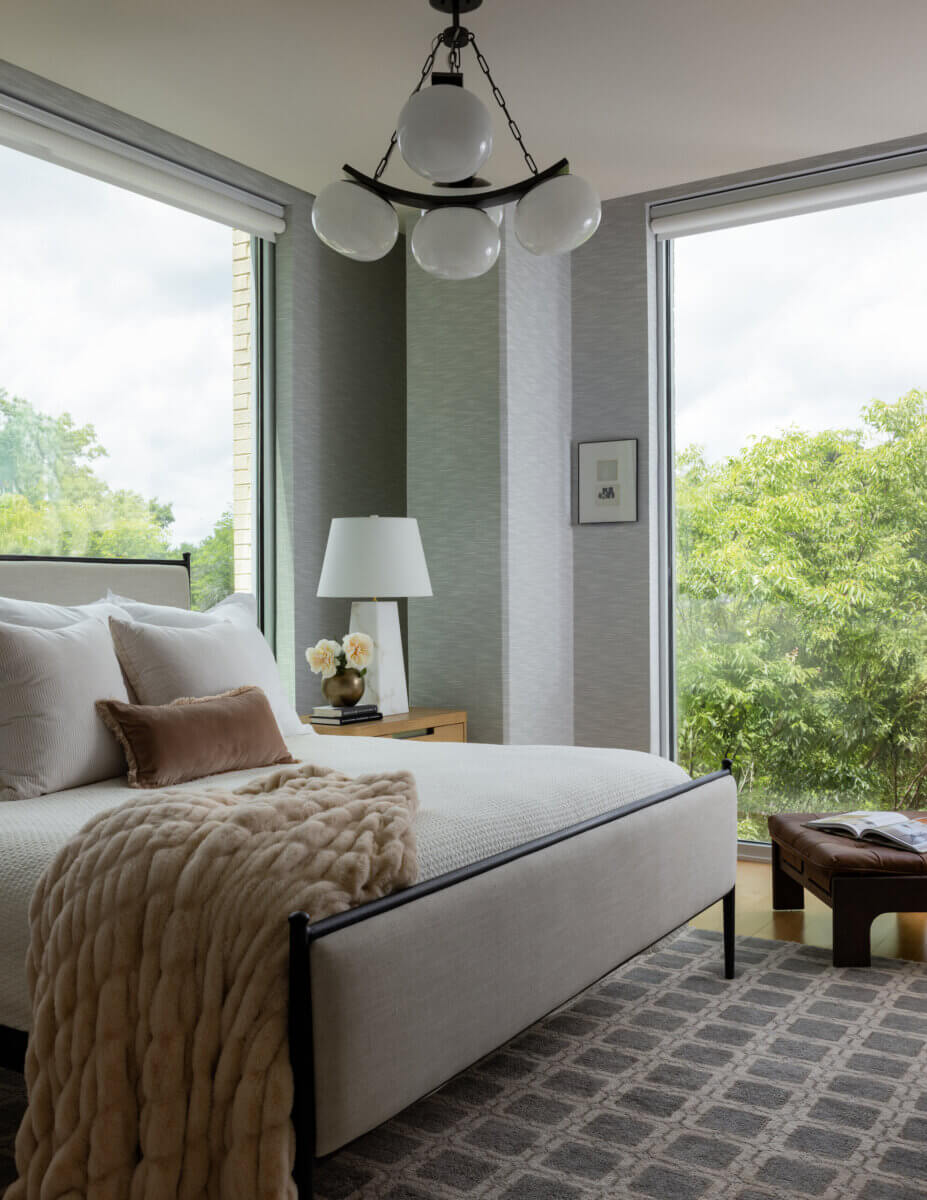 Whimsy Meets Cozy in This Dreamy Carolina Blue Home - 12 Modern bedroom with large windows, a beige upholstered bed, plush throw, bedside table with lamp and flowers, and a patterned rug. Greenery is visible outside.