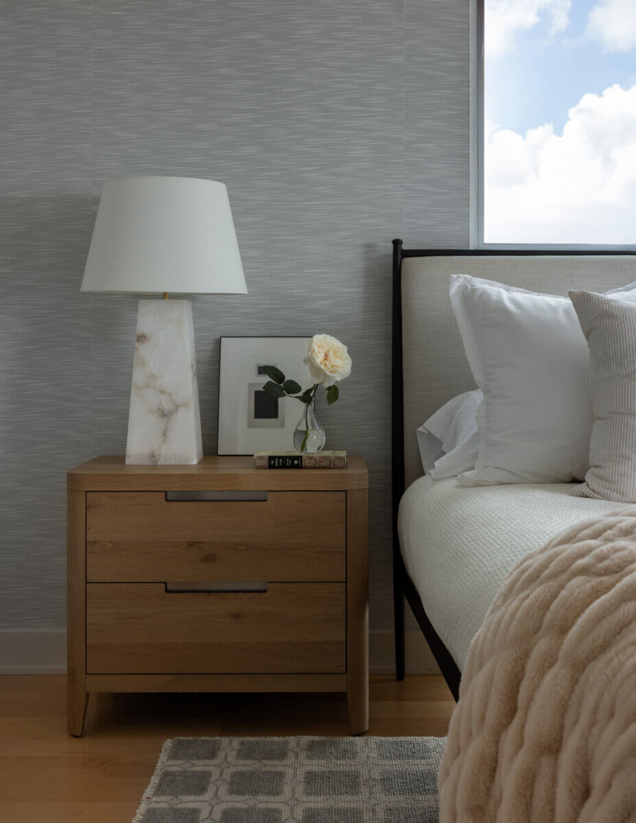 Whimsy Meets Cozy in This Dreamy Carolina Blue Home - 13 A wooden nightstand with a marble lamp, framed photo, book, and a vase holding a white rose next to a bed with white bedding and a beige throw. A window shows a cloudy sky.