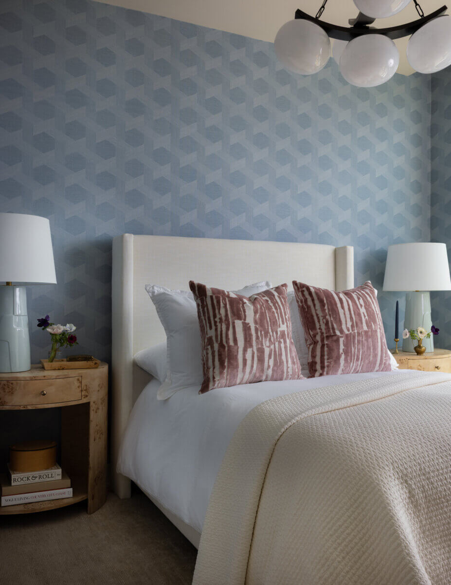 Whimsy Meets Cozy in This Dreamy Carolina Blue Home - 14 A neatly made bed with patterned pillows is flanked by two round wooden nightstands, each with a white lamp and small decor, set against a blue geometric wallpaper.