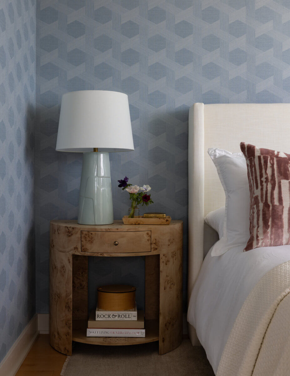 Whimsy Meets Cozy in This Dreamy Carolina Blue Home - 15 A wooden nightstand with books, a lamp, a small vase of flowers, and gold accents beside a bed with white and patterned pillows against a blue geometric wallpaper.