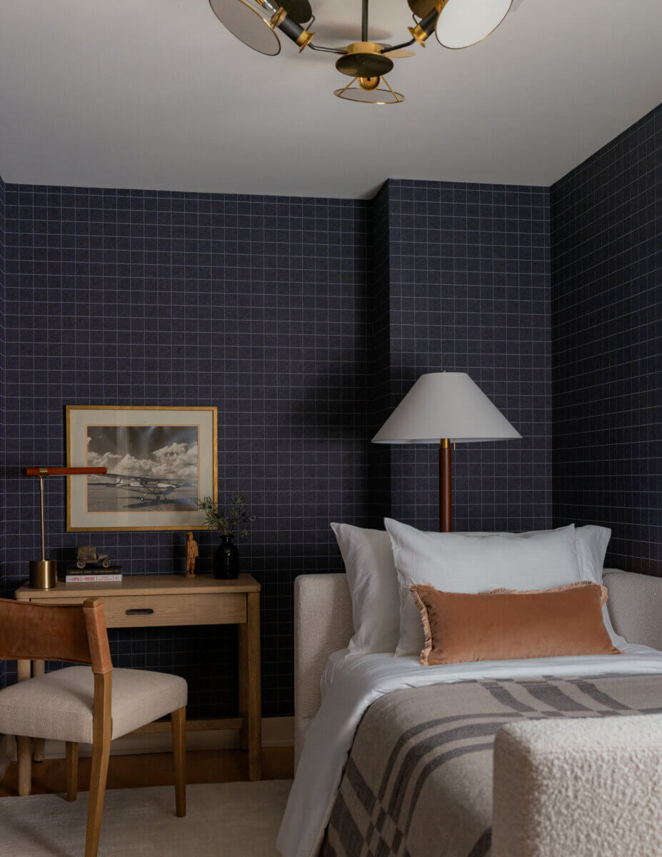 Whimsy Meets Cozy in This Dreamy Carolina Blue Home - 16 A small bedroom with a single bed, tan accent pillow, desk with chair, framed plane photo, table lamp, and dark grid-patterned wallpaper.
