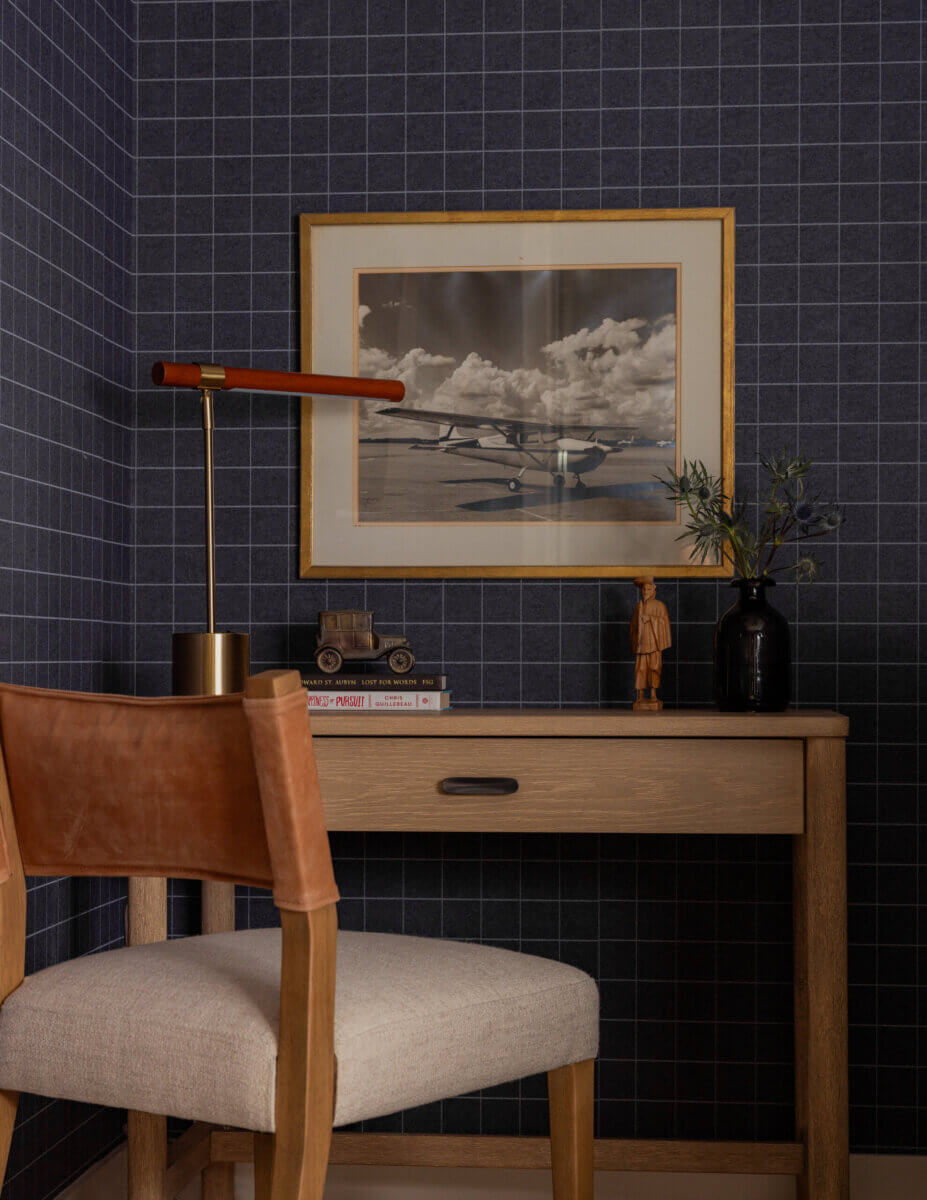 Whimsy Meets Cozy in This Dreamy Carolina Blue Home - 17 A small desk with a chair faces a framed airplane photo on a grid-patterned wall; a lamp, books, model car, figure, and vase decorate the desk.