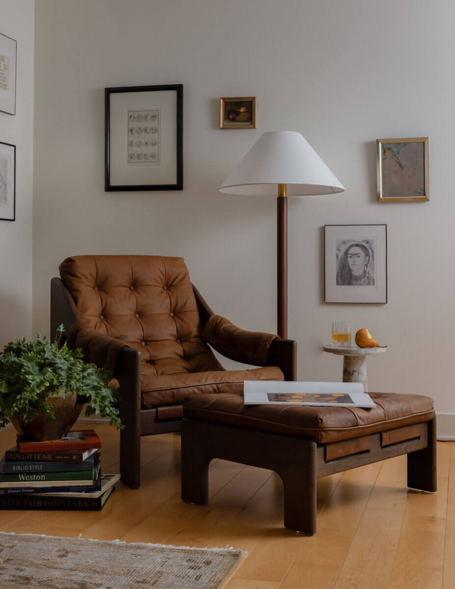Whimsy Meets Cozy in This Dreamy Carolina Blue Home - 11 A brown leather chair with matching ottoman sits in a corner, beside a floor lamp, framed art on the wall, a small table with books, a plant, a drink, and a plate with a pastry.