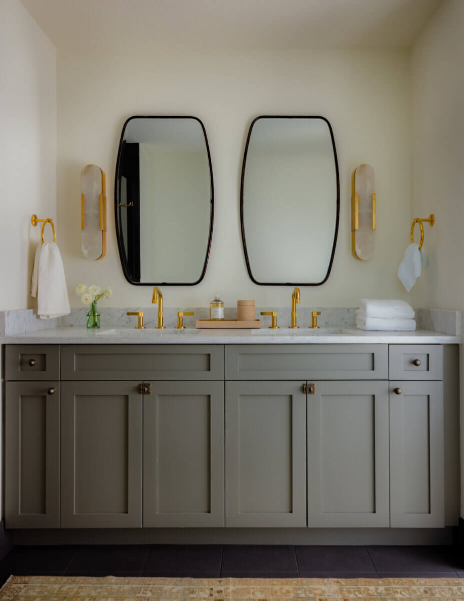 Whimsy Meets Cozy in This Dreamy Carolina Blue Home - 18 Modern bathroom with gray vanity, marble countertop, two mirrors, gold faucets, wall sconces, white towels, and a small vase of flowers.