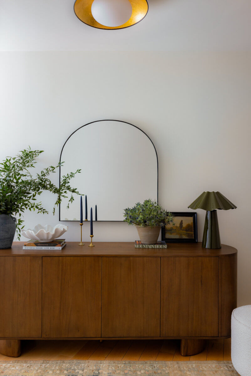 Whimsy Meets Cozy in This Dreamy Carolina Blue Home - 4 A wooden sideboard with plants, candles, books, a shell-shaped bowl, a table lamp, and an arched mirror against a white wall.