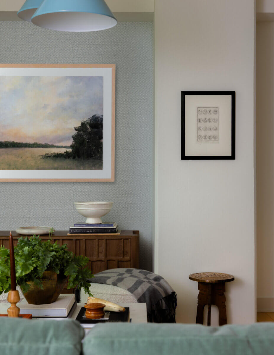 Whimsy Meets Cozy in This Dreamy Carolina Blue Home - 5 A living room corner with a wooden sideboard, books, plants, a ceramic bowl, framed art on the walls, a patterned blanket, a round stool, and a blue hanging light fixture.