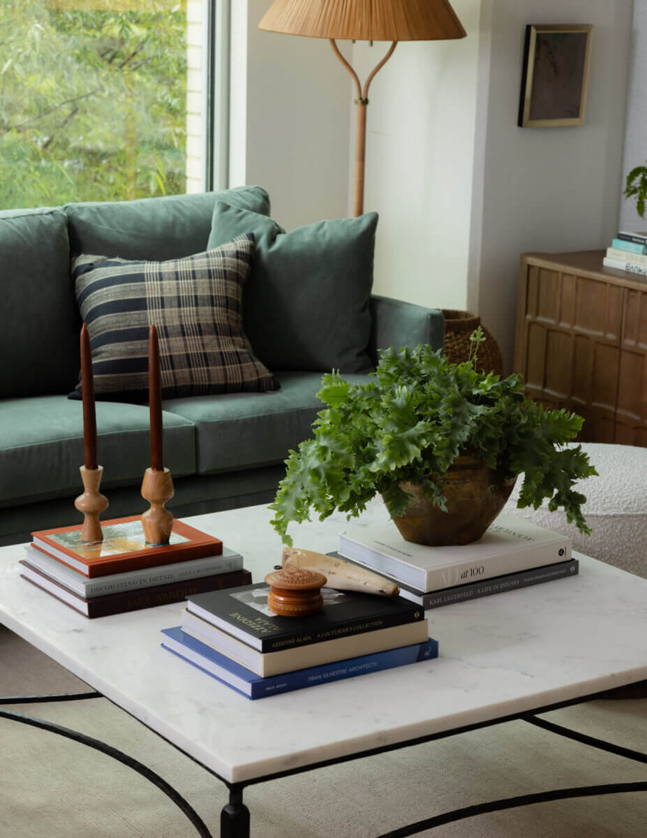 Whimsy Meets Cozy in This Dreamy Carolina Blue Home - 7 A marble coffee table with stacked books, wooden candlesticks, a potted green plant, and a small round box sits in front of a green couch with a plaid pillow.