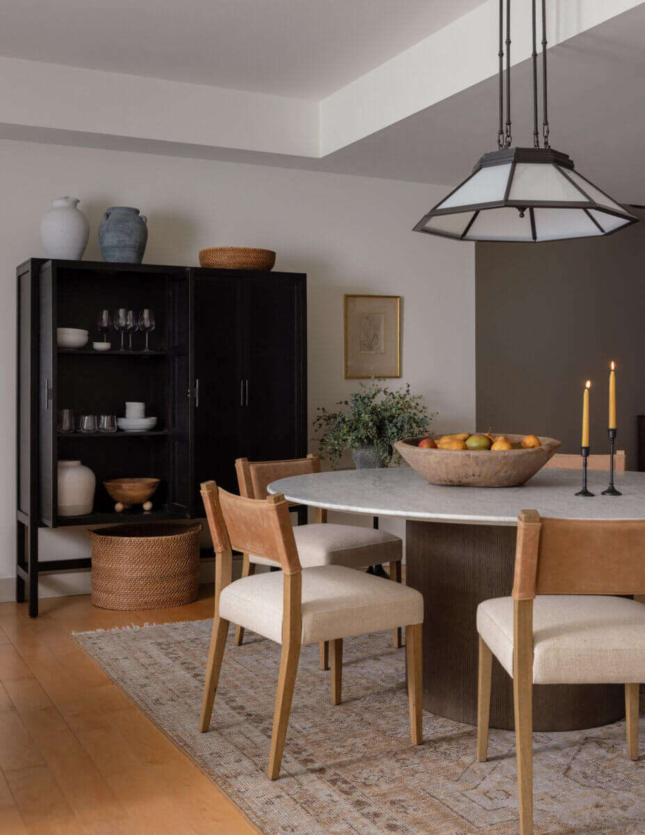 Whimsy Meets Cozy in This Dreamy Carolina Blue Home - 9 Modern dining room with a round table, four chairs, two lit candles, a fruit bowl, a rug, and a black cabinet with ceramics and glassware against a white wall.