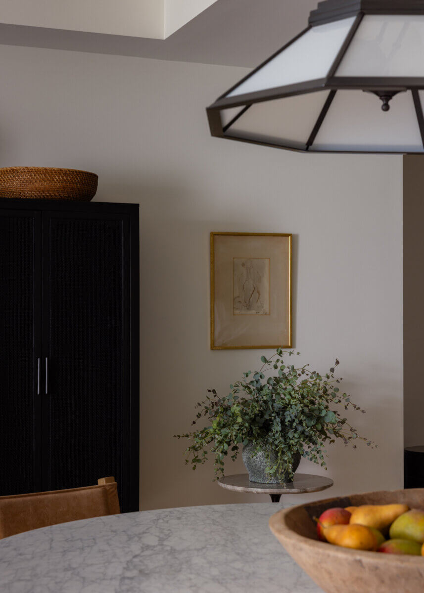 Whimsy Meets Cozy in This Dreamy Carolina Blue Home - 10 A round marble table with a bowl of fruit sits beside a potted plant on a small stand, a black cabinet, and framed art on a beige wall under a geometric ceiling light.