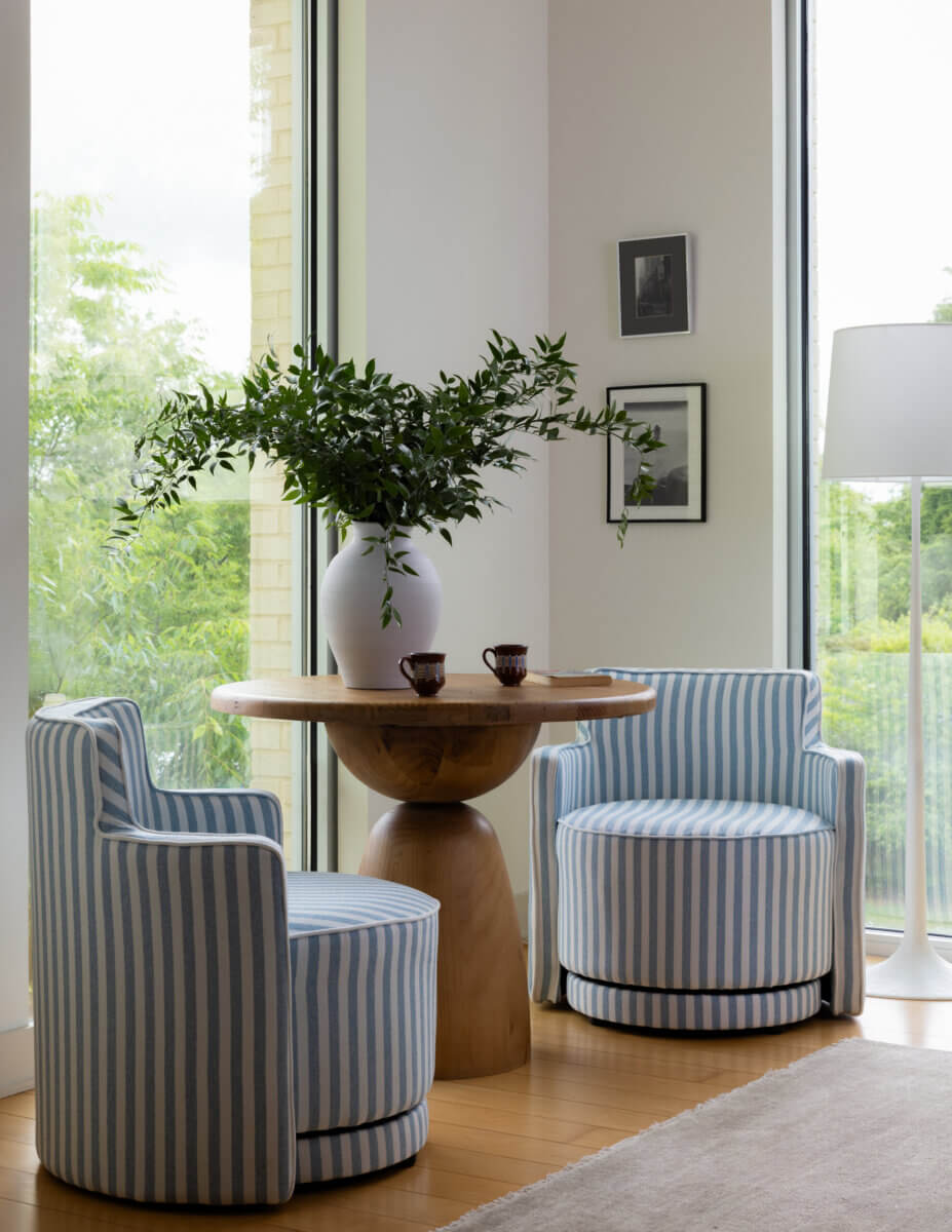 Whimsy Meets Cozy in This Dreamy Carolina Blue Home - 8 Two blue-and-white striped chairs sit by a round wooden table with a white vase of greenery, near large windows and a white floor lamp.