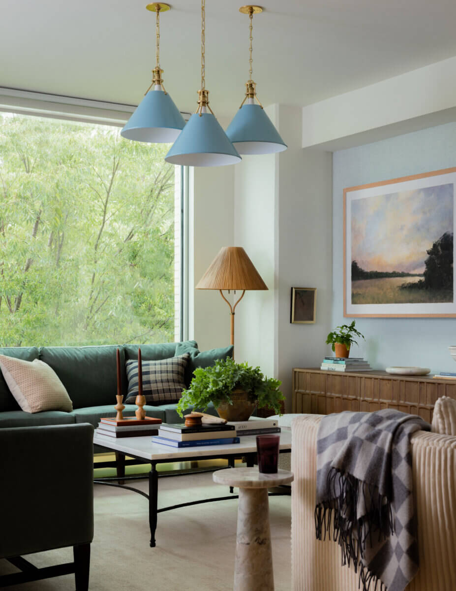 Whimsy Meets Cozy in This Dreamy Carolina Blue Home - 6 Modern living room with green sofa, coffee table with books and plants, plaid throw blanket, large window, pendant lights, and landscape painting on the wall.