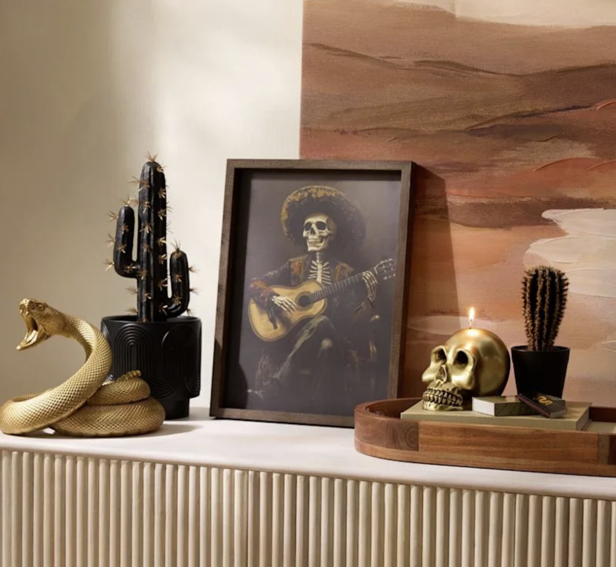20 Halloween Decor FINDS That Are Actually Chic - 10 A framed skeleton guitarist print with gold snake sculpture, gold skull candle, and two potted cacti are displayed on a beige surface in front of abstract wall art—perfect Halloween decor to elevate your space this spooky season.