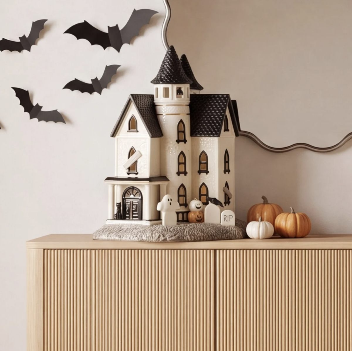 20 Halloween Decor FINDS That Are Actually Chic - 16 A decorative haunted house, small pumpkins, ghost figurines, and bat wall decals sit on a wooden cabinet next to a mirror, creating the perfect Halloween decor for the spooky season.