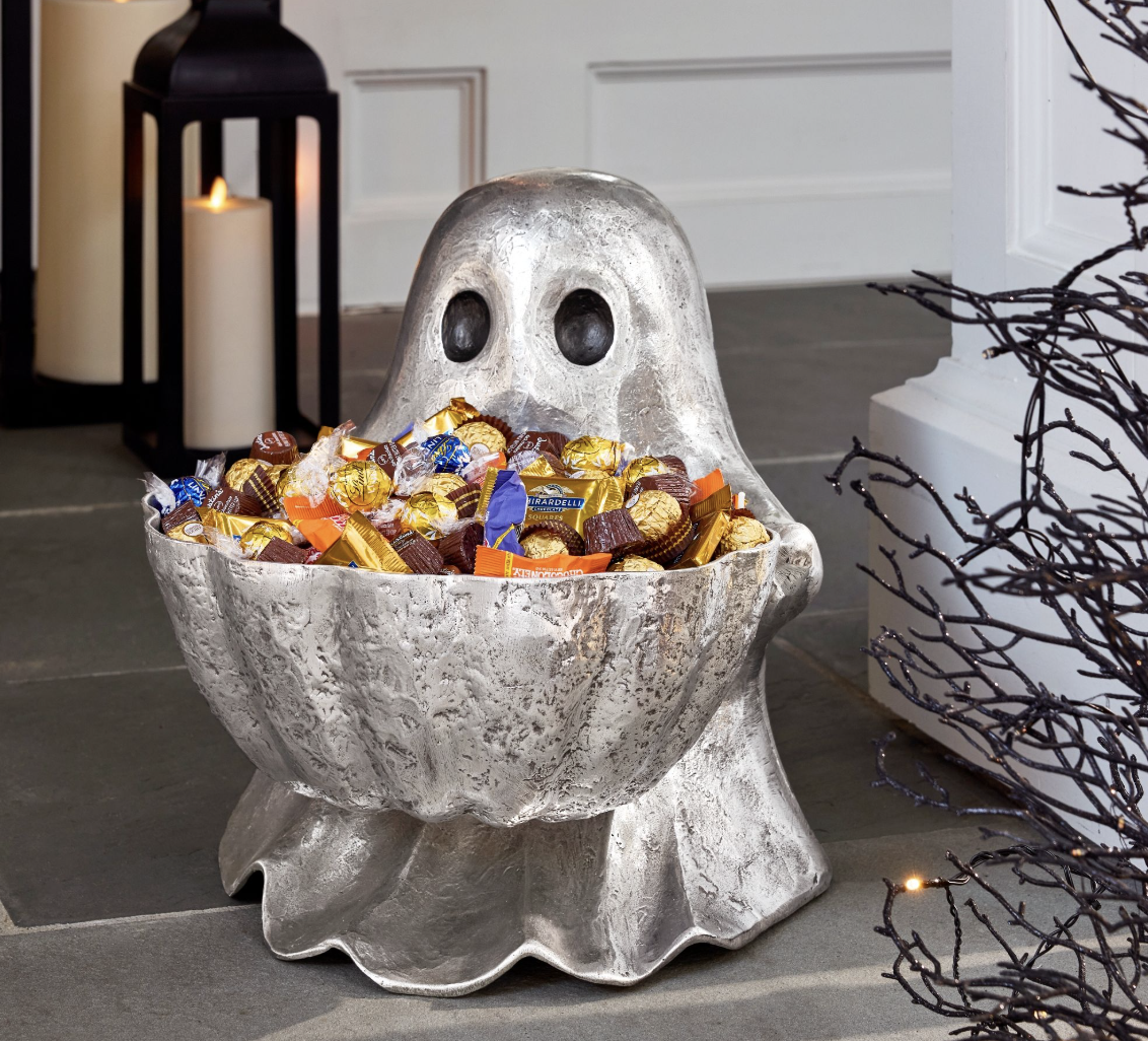 20 Halloween Decor FINDS That Are Actually Chic - 14 A silver ghost-shaped bowl filled with assorted wrapped candy sits on a porch next to candles and a bare decorative branch—an eye-catching piece of Halloween decor perfect for welcoming guests during Spooky Season.