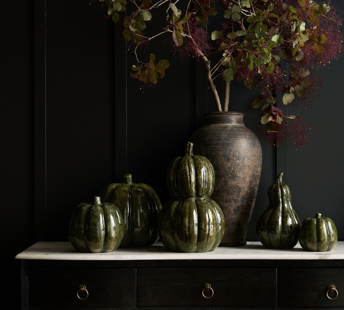 20 Halloween Decor FINDS That Are Actually Chic - 18 Six glossy green ceramic gourds are arranged on a dark wooden dresser beside a tall brown vase filled with dark red leafy branches, set against a black paneled wall—an elegant display of Halloween decor perfect for the spooky season.