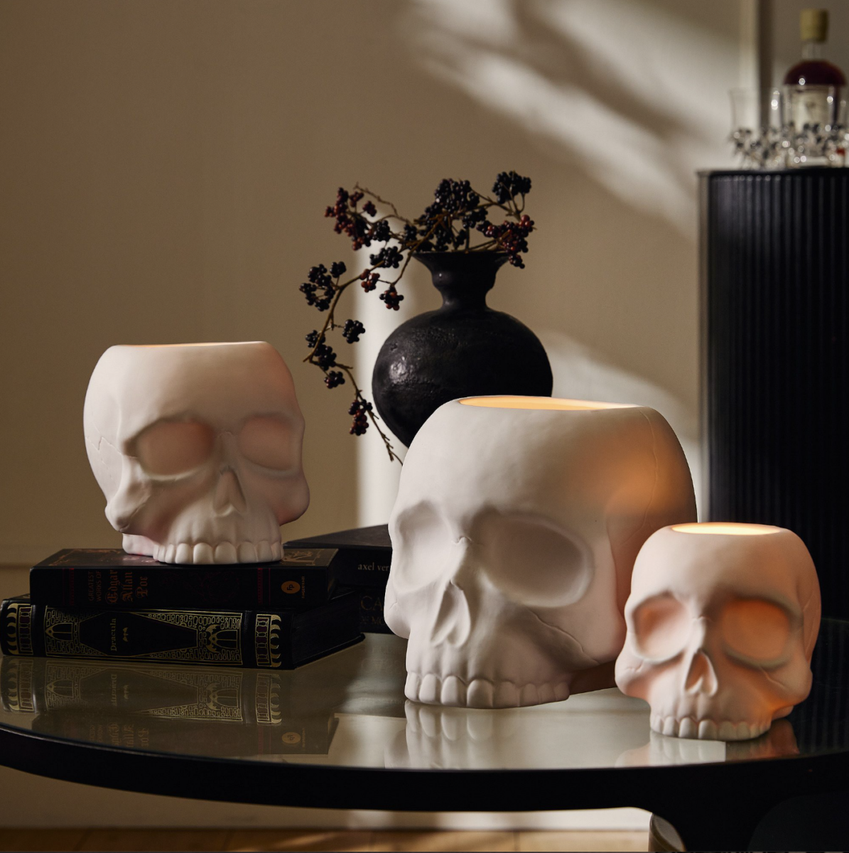 20 Halloween Decor FINDS That Are Actually Chic - 15 Three white skull-shaped candle holders sit on books and a glass table, adding the perfect touch of Halloween decor; a black vase with branches decorates the background for an effortlessly spooky season display.